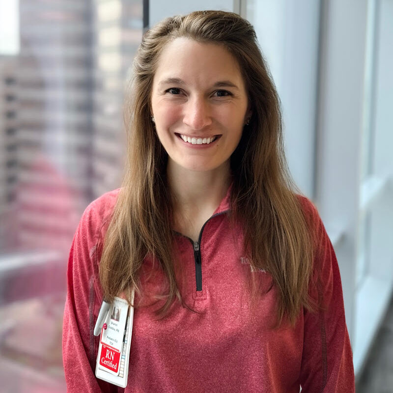 A woman with long brown hair wearing a red zip-up top and an RN badge stands smiling by a window with city buildings in the background.