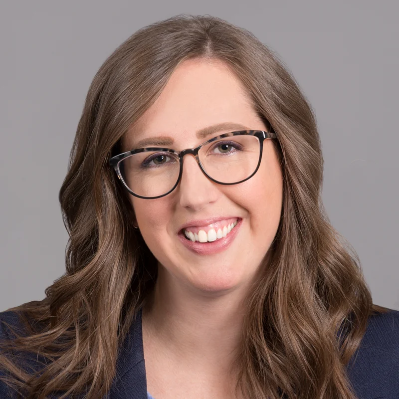 Woman with long brown hair and glasses, wearing a navy blazer and light blue blouse, smiling in front of a neutral gray background.