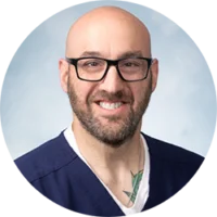 A smiling man with a shaved head, glasses, and a dark blue scrub top.